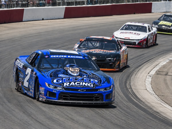 Grumpy Old Geezer Racing stock car leading several cars through a paved oval-track corner